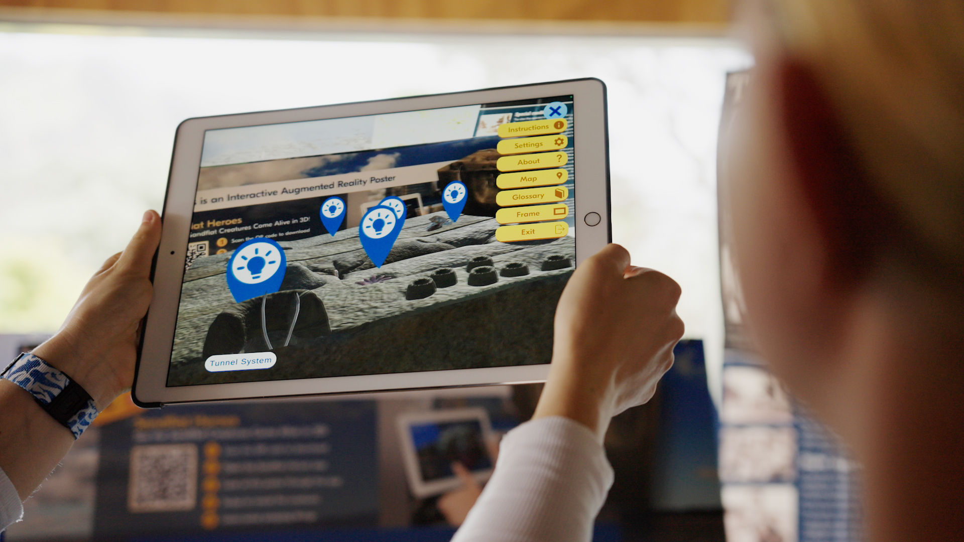 A person holding a tablet displaying the Sandflat Heroes AR game overview map with multiple creature location markers visible on screen