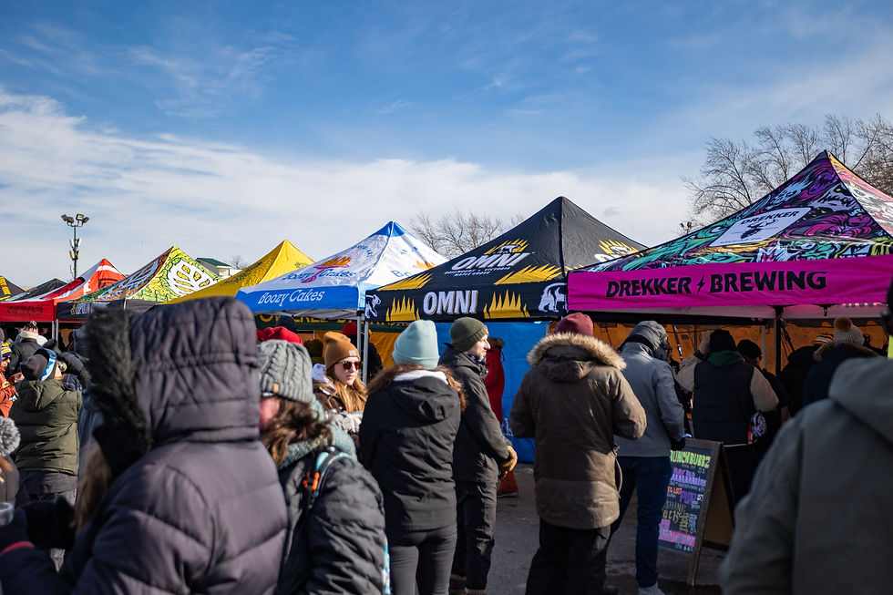 Outdoor market scene with people in winter coats, browsing colorful tents labeled OMNI and DREKKER BREWING. Clear sky, lively atmosphere.