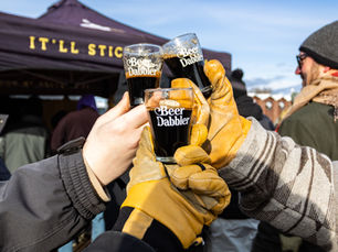 3 hands with yellow leather gloves on, clinking 3 small glasses full of dark liquid that say "Beer Dabbler"