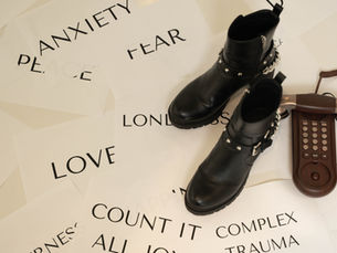 Black ankle boots with silver chains stand beside a vintage brown telephone on a white floor scattered with words including “anxiety,” “fear,” “love,” “loneliness,” and “complex trauma.”