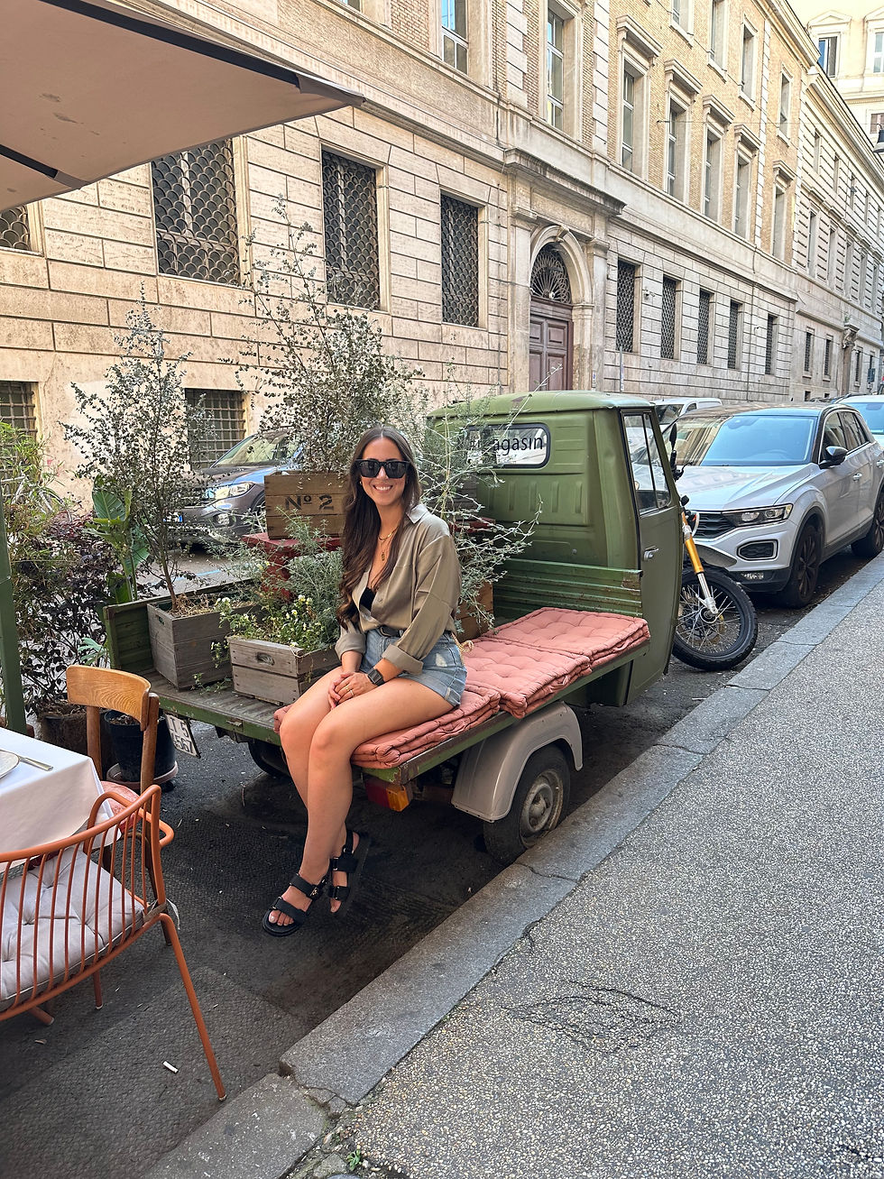 A female with sunglasses sitting on the bed of a truck next to flowers on the streets of Rome
