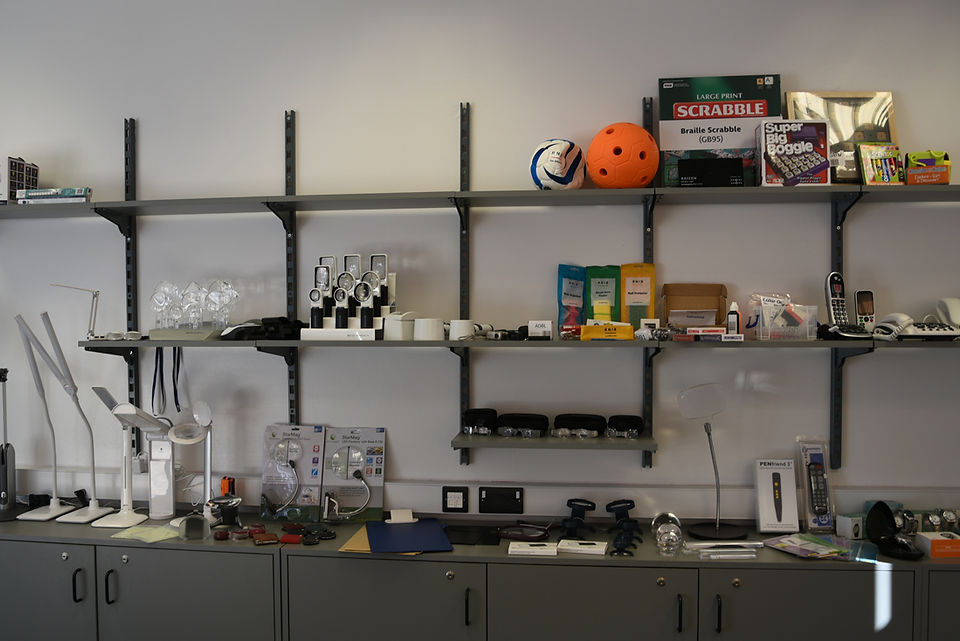 A wide angle photograph of some of the accessible items we have on display. This includes; a football and games, on the top shelf; magnifiers on the left of the middle shelf, and various items such as sock organisers on the right, and lamps and digital resources on the bottom shelf.