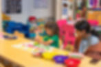 Student painting at a classroom table while a teacher looks on during a creative activity.