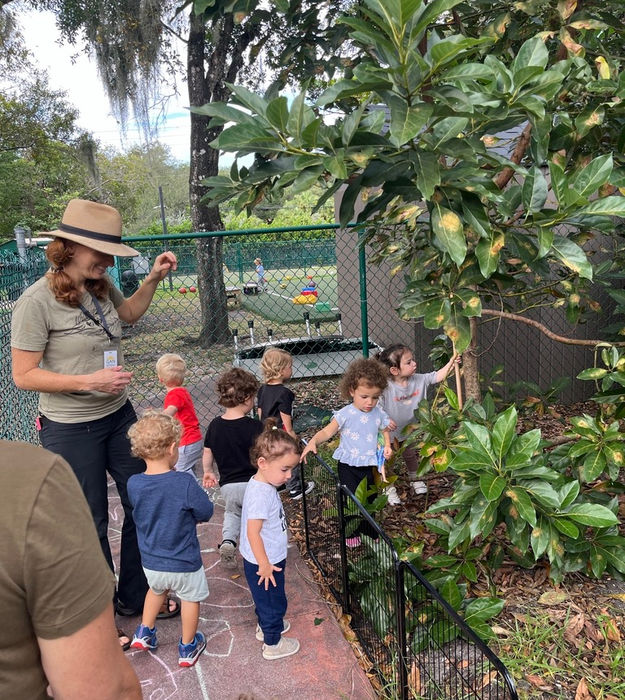 Preschool teacher leading young children on a nature walk through a garden path during an outdoor learning activity.