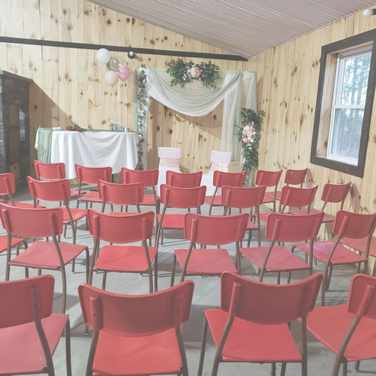 Salle aménagée pour une cérémonie avec des rangées de chaises rouges alignées face à une décoration florale composée de voilage blanc, de guirlandes vertes et de touches de rose, parfaite pour un mariage ou une célébration intime.
