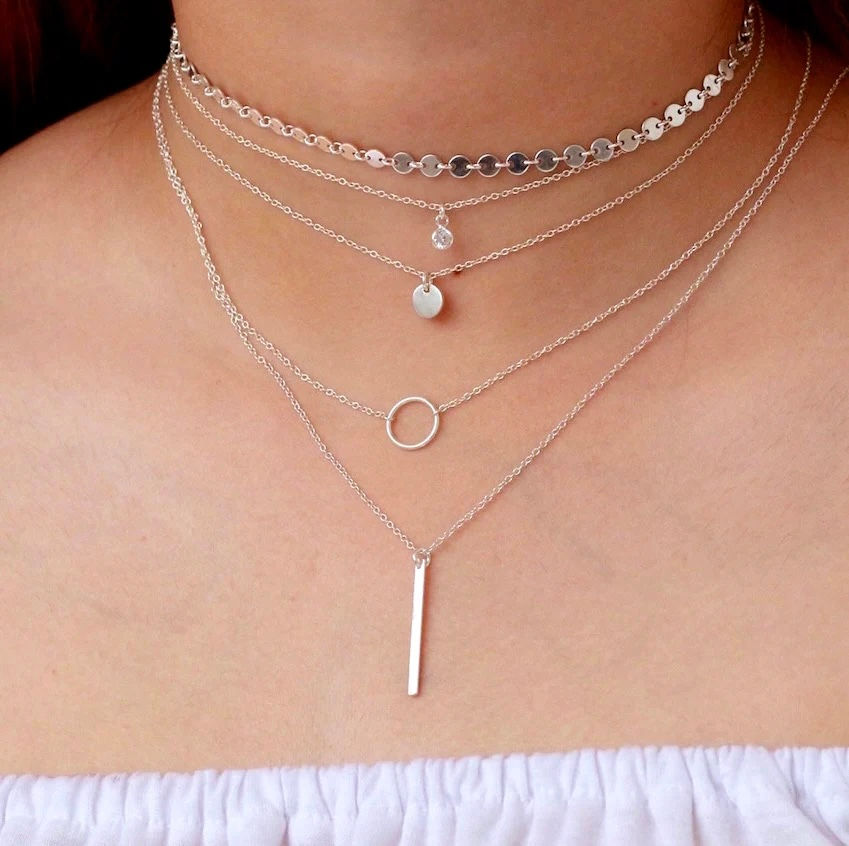 A close-up of a woman’s neck adorned with layered silver necklaces, including a delicate choker, minimalist pendants, and fine chain details. The polished silver tones create a sleek and modern aesthetic, styled against a soft, neutral off-the-shoulder outfit for an effortlessly chic look.