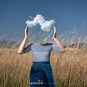 A person standing in a field holds up a circular mirror where their face would be, reflecting a clouded blue sky—symbolising identity, self-reflection, and change.