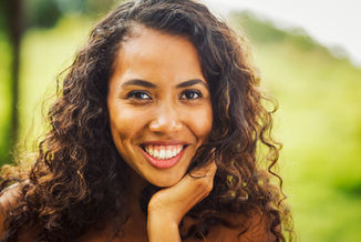 A black woman with dark brown curly hair resting her head in her hand smiling