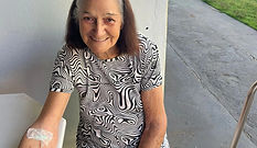 Idosa sorrindo sentada em uma cadeira, com curativo no braço, demonstrando conforto e recuperação no ambiente externo do residencial Cura D’ars.