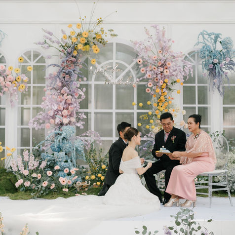 The bride and groom seated on a white stage during an outdoor wedding ceremony, flanked by family members.