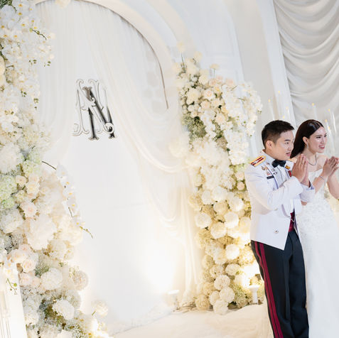 A candid moment on stage as the groom leans in to speak with his smiling bride amidst white rose decorations.