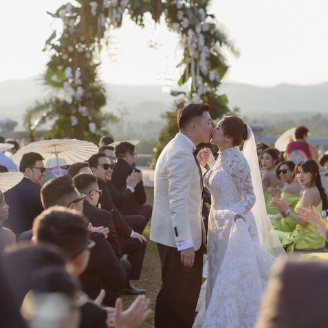 The bride and groom sharing a romantic first kiss at the altar under a lush green floral arch with a mountain view backdrop.