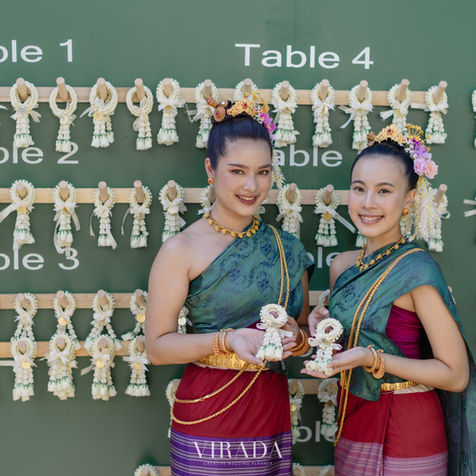 Two beautiful hostesses dressed in traditional dark green and crimson Thai silk wraps smiling by a guest check-in wall.