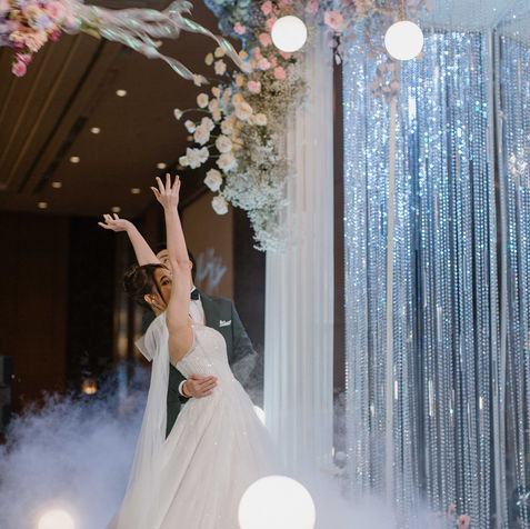 The bride in a white gown joyfully tossing her bouquet on a stage filled with low-lying fog.