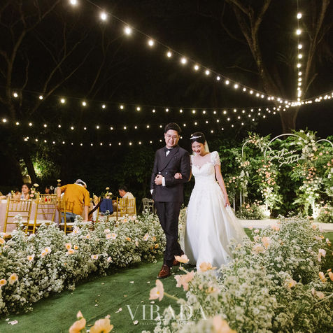 The couple walking together along a path lined with flowers under string lights.