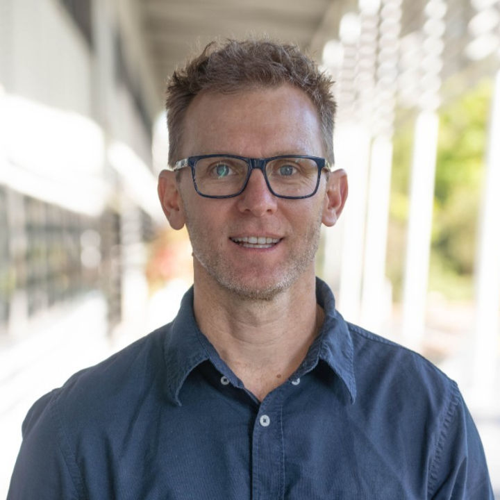 Man with glasses and a blue shirt stands smiling in a bright outdoor setting with a blurred background of a walkway and greenery. Sports scientist, Scott McLean.