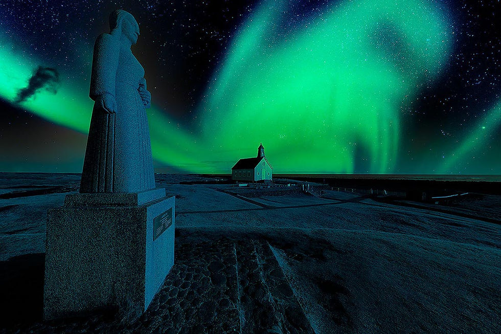 Statue in foreground under vivid green aurora borealis. A small illuminated church sits in the distance against a starry night sky.