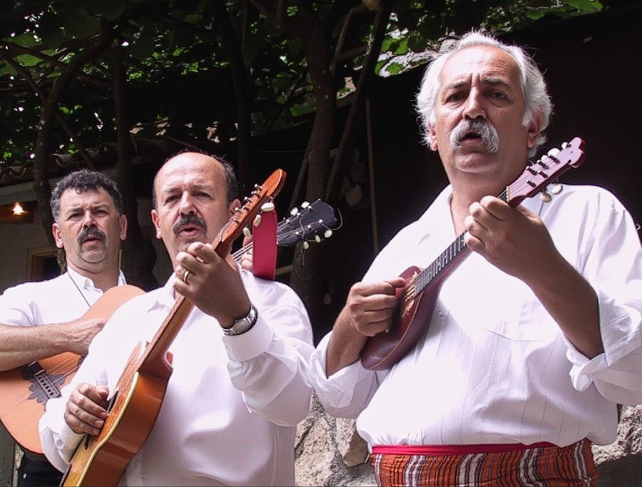Three men in white shirts play string instruments, singing outdoors under leafy trees. The mood is lively and engaged.