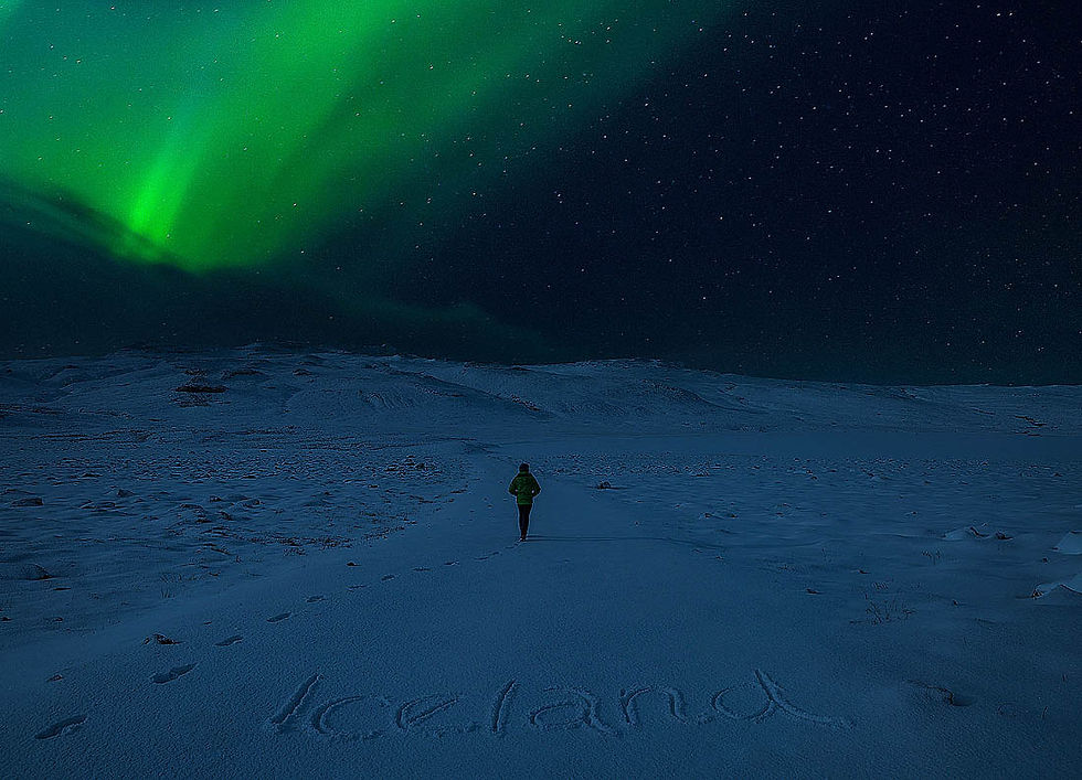 Person in a green jacket walks on snowy terrain under vibrant green aurora, with "Iceland" written in the snow, starry night sky above. Matjaz Intihar, fotopotep potovanje, Severni sij.