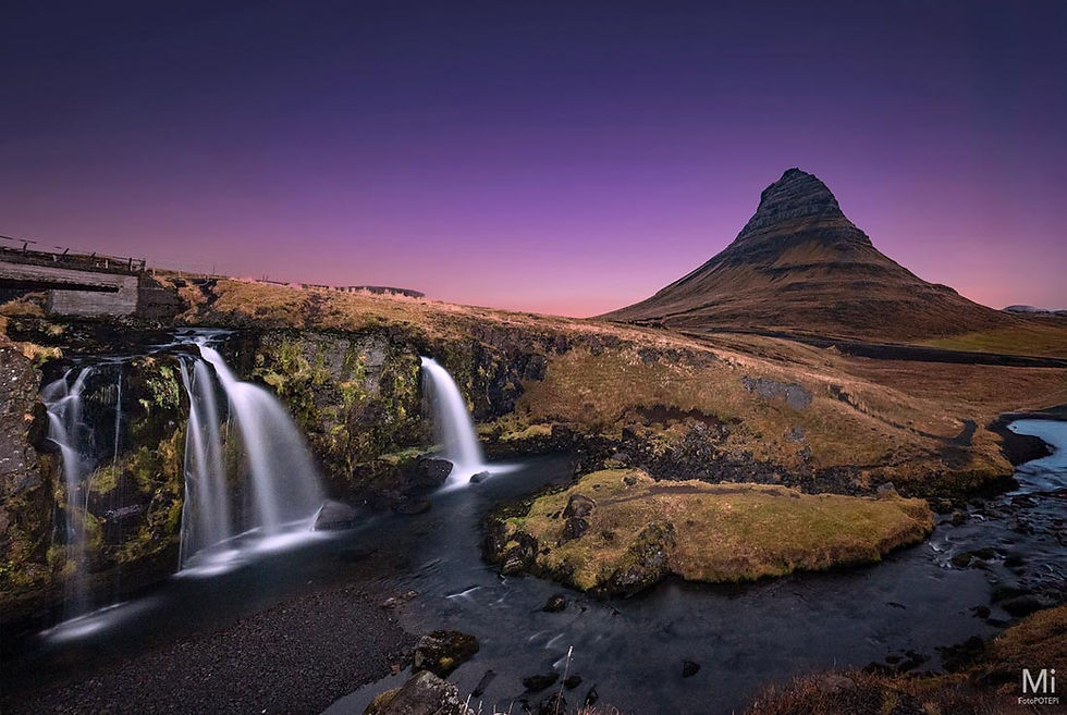 Kirkjufell mountain at sunset, reflecting in still water with nearby waterfall in foreground. Dramatic sky over Iceland’s iconic peak.