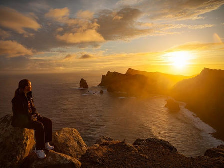 Popotnica sedi na skali in opazuje zlati sončni zahod nad razgibano obalo Madeire.