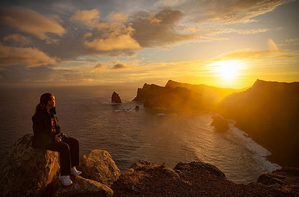 Person sitting on a rock, gazing at a golden sunset over a rugged coastline. The sky is partly cloudy with warm hues, creating a serene mood. Madeira, Portugal, Matjaz Intihar, FotoPotep.