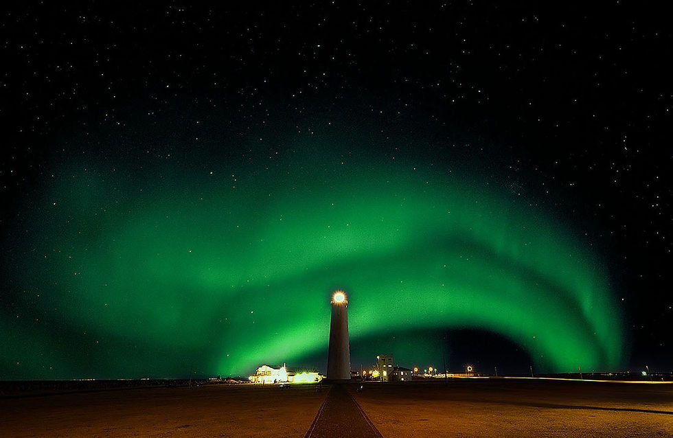 A lighthouse under vivid green northern lights in a starry night sky. The setting is serene, with buildings faintly illuminated below. Matjaz Intihar, fotopotep potovanje, Severni sij.