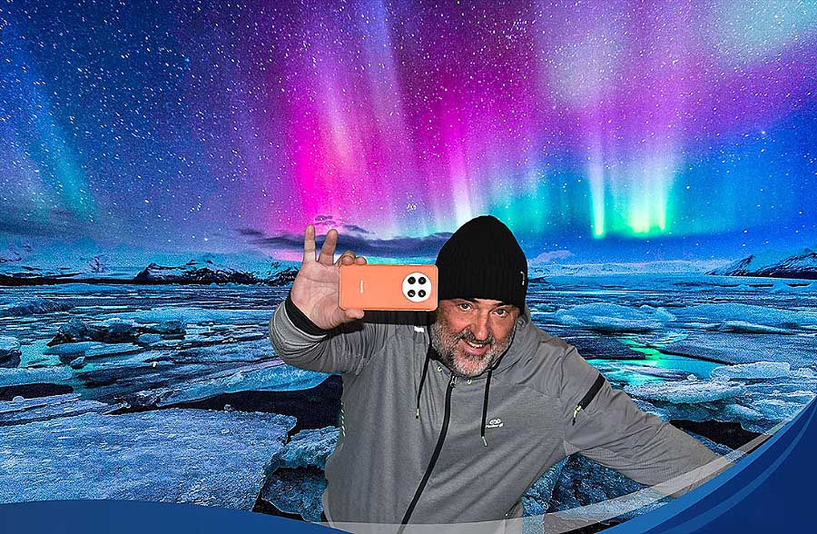 Man in gray jacket taking a selfie with an orange phone under vibrant Northern Lights. Icy landscape in background, wearing a black beanie.