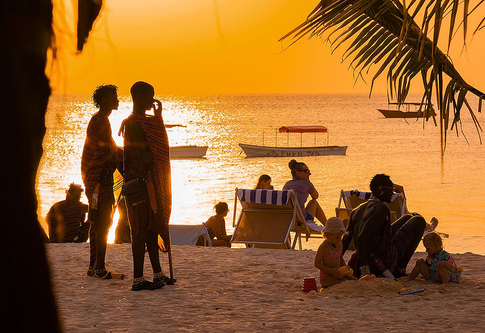 People enjoy a sunset on a beach, with some seated on chairs and others standing. Children play in the sand. The sky and sea glow orange.