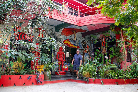 Man in blue shirt stands in front of colorful, mural-covered building with plants and red accents. Signs for restaurant and bar visible.