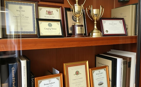 8 or 9 certificate style and cup awards on 2 wooden shelves in an otherwise full bookcase.