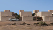 Architectural visualisation of the street elevation of a contemporary Emirati residence designed by TMD Studio. The façade presents a sequence of monolithic sand-toned volumes with curved corners and recessed entrances, creating a protective street edge while framing moments of transition into private gardens. Vertical ventilation shafts rise above the roofline, each perforated with patterned mashrabiya openings for airflow and light filtration. Palm trees and native vegetation soften the courtyard interiors, only partly visible from the street. A white SUV passes in front, emphasising scale and the house’s relationship to the desert road. The design illustrates cultural privacy, climatic resilience, and the reinterpretation of traditional Emirati thresholds within a modern architectural language.