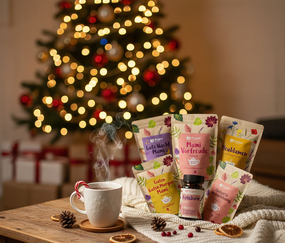 Weihnachtliches Arrangement mit Bio-Tees für Schwangerschaft, Folsäure und Heublumen-Sitzbad auf einem Holztisch vor einem beleuchteten Weihnachtsbaum, daneben eine dampfende Tasse Tee.