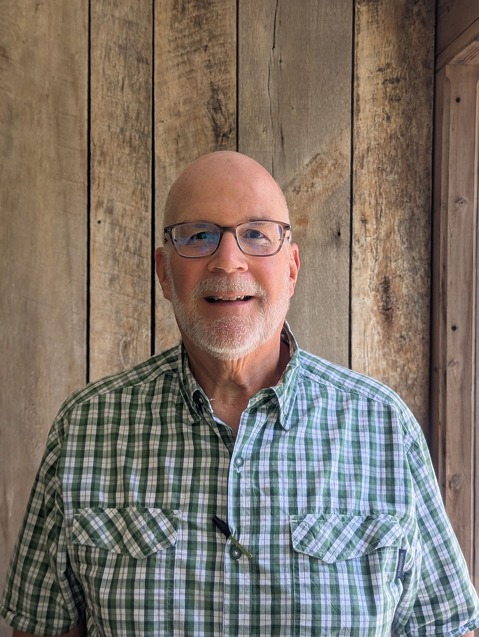 Portrait photo of a smiling older adult wearing glasses and a green plaid shirt, standing in front of rustic wooden boards, portrait orientation.