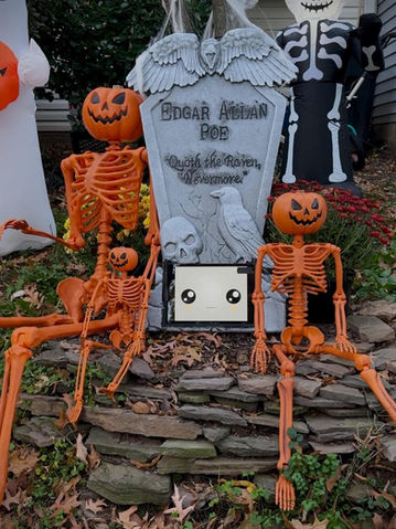 A headstone with a tablet at the bottom next to two orange skeletons with pumpkins as heads. Another skeleton is in the back.