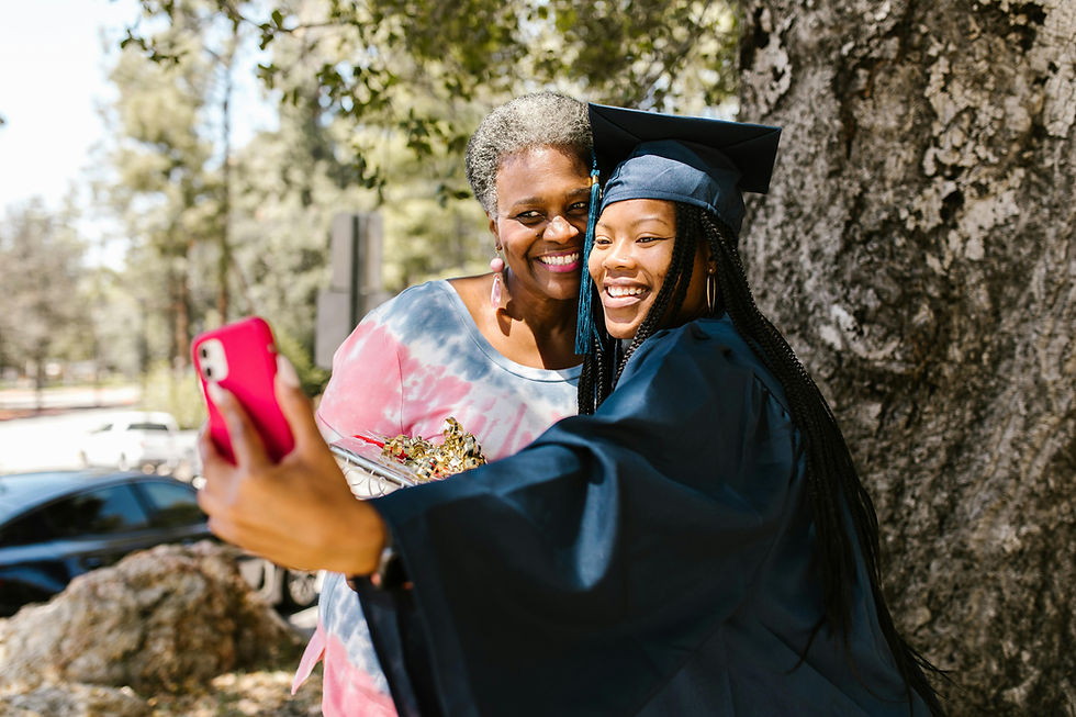 Graduate taking selfie with mother, smiling brightly near a large tree.