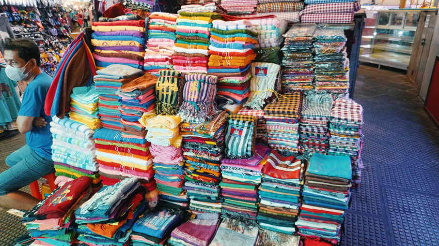 Serviettes, sarongs et krama traditionnels s'empilent en montagnes colorées dans l'une des allées textiles du marché, sous l'œil discret d'un vendeur masqué