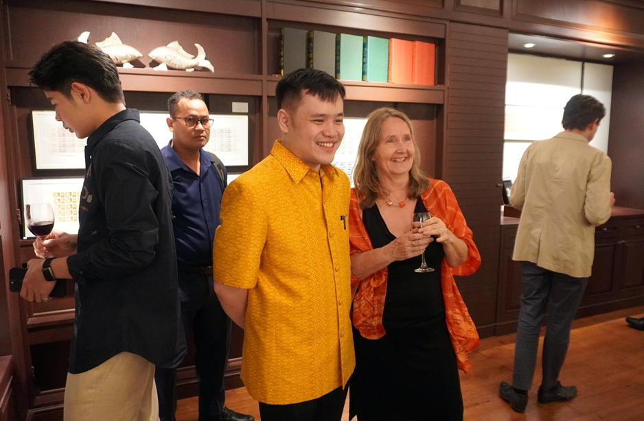 Sereyvong Jason Chhinlim, in a yellow silk shirt, poses with a guest in front of framed stamp sheets at the opening