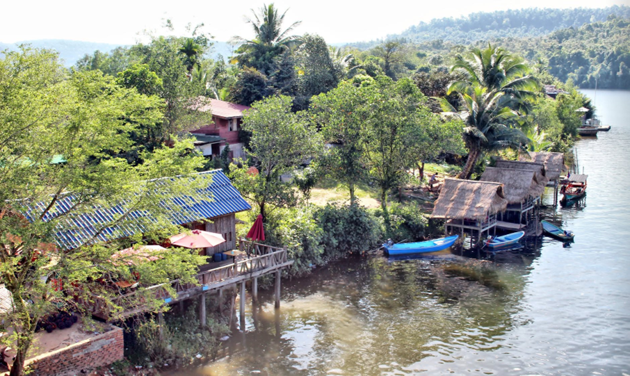 Vue plongeante sur le village riverain : toits de tôle bleue et toits de chaume se côtoient au bord de l'eau brune, sous une canopée continue qui rappelle la proximité immédiate de la forêt des Cardamomes
