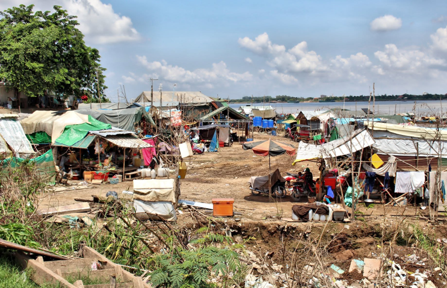 Vue d'ensemble du campement : tentes de bâches, cuisines en plein air et motos garées entre les abris de fortune, avec le fleuve et la ville en toile de fond