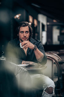 junger mann mit kaffeetasse in der hand beobachtet das stadtgeschehen aus von einem strassekaffee aus, aufgenommen von renee art photography