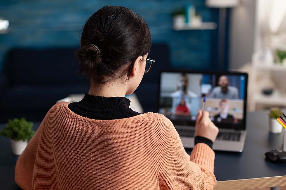 Person sits at a desk on a laptop video call with multiple participants on screen, holding a pen as if taking notes.