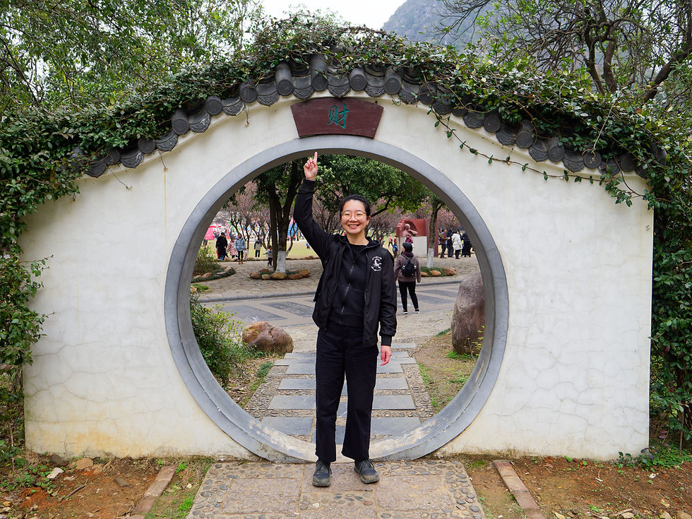 A Chinese woman standing in front of a door with a decorative word "fortune". She points at the door, smiling. She's wearing a black jacket and black pants.