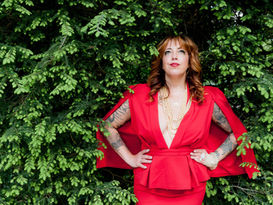 Rose Kaz, in a red cape-dress, looking up. She is standing in front of a green bush.