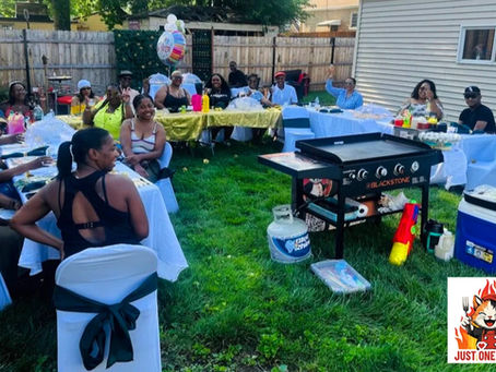 Group of friends at a backyard cookout seated at white-covered tables around a Blackstone grill; Just One Hibachi logo visible.