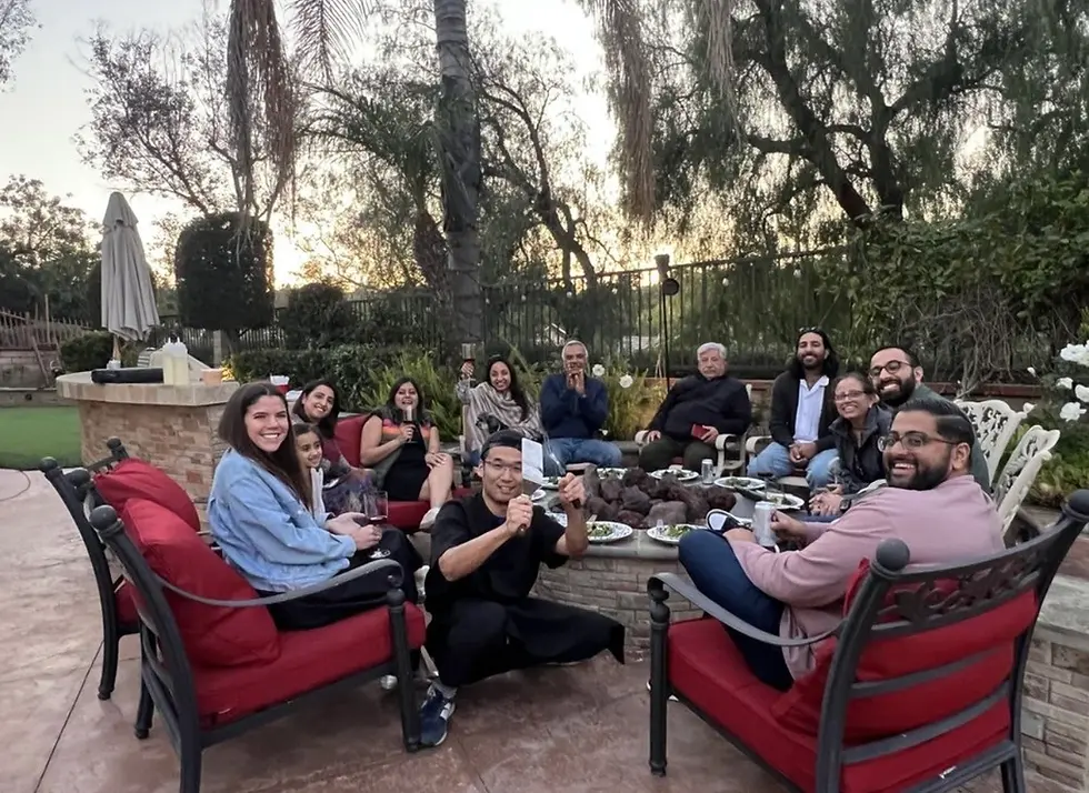 Smiling group enjoys food around a fire pit in a backyard patio. Red chairs and greenery enhance the relaxed, cheerful atmosphere.