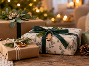 Weihnachtlich verpackte Geschenke in Kraftpapier und dunkelgrünem Geschenkpapier mit Schleifen, Tannenzweigen, Tannenzapfen und getrockneten Orangenscheiben auf einem Holztisch. Kerzenlicht und ein beleuchteter Weihnachtsbaum sorgen im Hintergrund für eine gemütliche, festliche Atmosphäre.