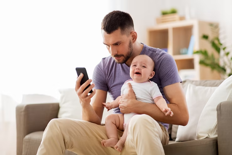 A man in a purple shirt sits on a sofa, looking at his phone while holding a crying baby. Bright, soft-focus living room setting.