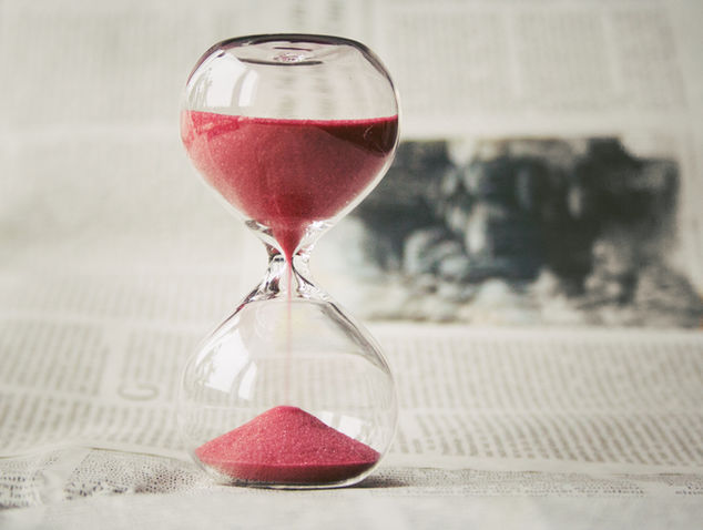 An elegant hourglass with vibrant pink sand slowly marks the passage of time against the backdrop of a blurred newspaper page.