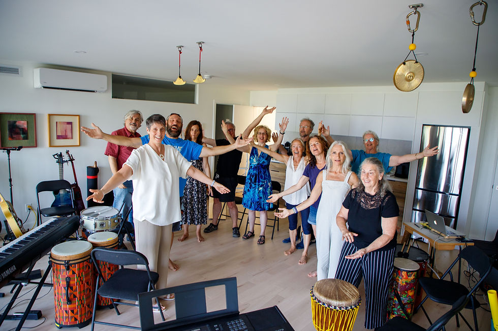 Group of people joyfully posing with arms outstretched in a music room with drums, keyboard, and framed artwork on walls.